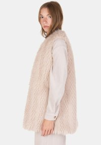 Chaleco de piel sintética en rosa claro, con superficie texturizada y diseño de frente abierto, combinado con una camiseta blanca de manga larga acanalada.