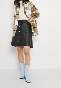 Kvinna iklädd en beige polotröja, brun rutigt jacka, svart knapp-up läderkjol och ljusblå ankellånga stövlar, stående mot en vit vägg.