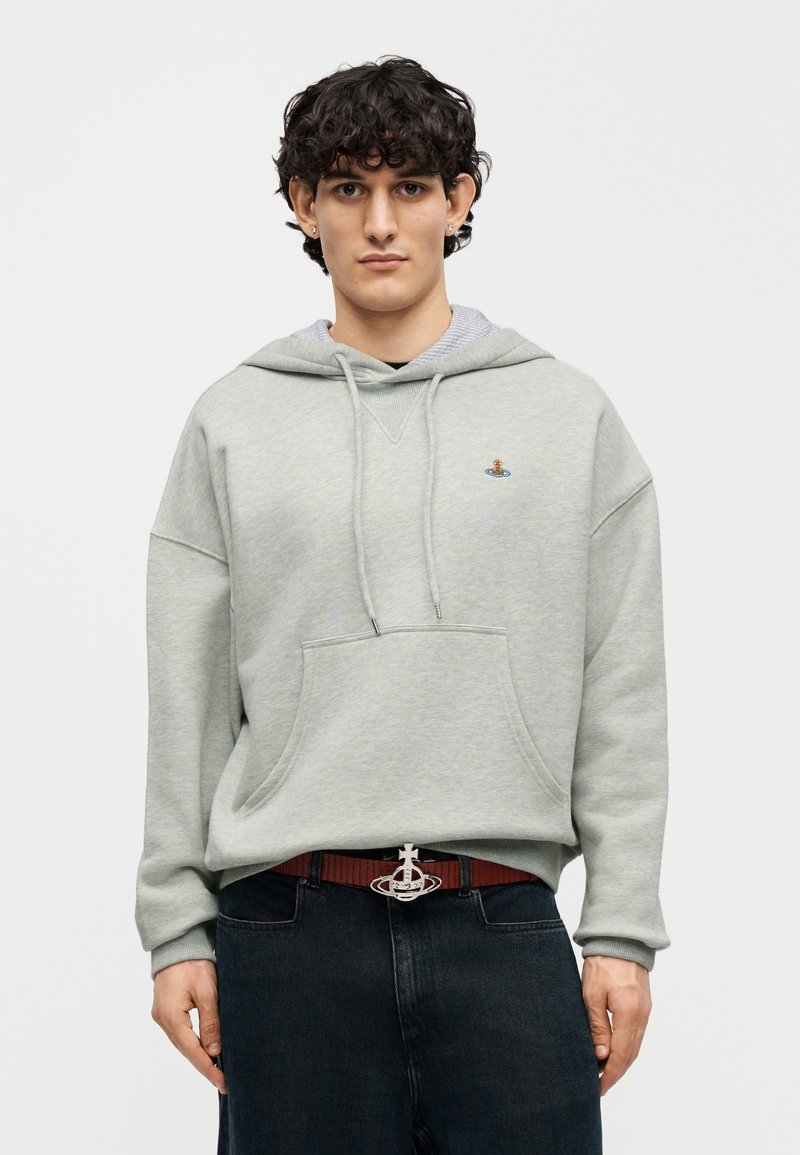 Sweat à capuche gris clair avec une poche avant, une capuche à cordon et un logo brodé. Porté avec un jean foncé et une boucle de ceinture argentée.