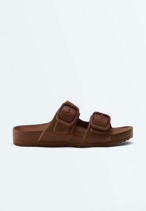 Brun slip-på-sandal med to justerbare spænderemme og en tekstureret sål, vist fra siden mod en ensfarvet baggrund.