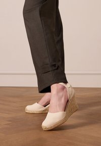 Wedge espadrille in licht crèmekleurige canvas met een geweven achterkant, touwzool en decoratieve strikaccent. Gecombineerd met donkere, op maat gemaakte broek.