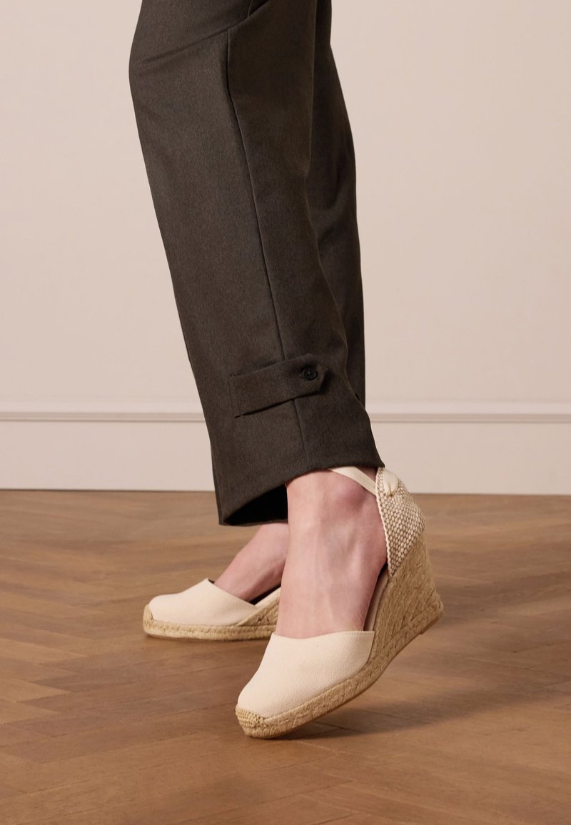 Wedge espadrille in licht crèmekleurige canvas met een geweven achterkant, touwzool en decoratieve strikaccent. Gecombineerd met donkere, op maat gemaakte broek.