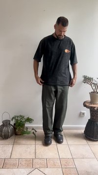 T-shirt preta de algodão com gráfico colorido de sol, corte oversized. Calças verde-oliva, de perna reta, combinadas com sapatos pretos. Ambiente interior.