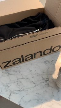 Juodas džemperis dalinai matomas atidaro Zalando kartoninėje dėžėje. Dėžėje yra ryškūs juodi užrašai ant rudos paviršiaus.