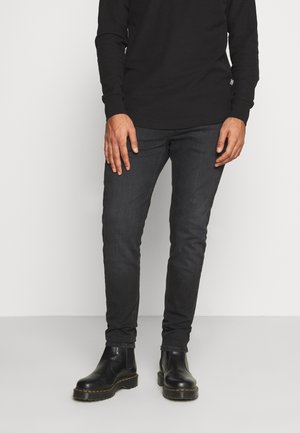 Homme portant un jean slim noir, une chemise noire à manches longues et des bottes en cuir noires, debout devant un fond clair uni.