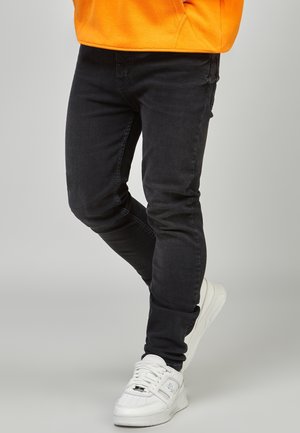 Person trägt schlanke schwarze Jeans, weiße Sneakers und ein oranges Sweatshirt vor einem schlichten hellen Hintergrund.