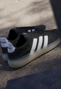 Sneakers Adidas nere con strisce bianche e suole traslucide posate su terreno in cemento alla luce soffusa del sole.