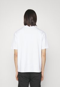 Bílá krátkosrstá tričko s kulatým výstřihem, vyrobené z hladkého materiálu, zobrazené zezadu, v kombinaci s tmavými cargo kalhotami.