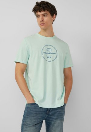 Junger Mann mit lockigem Haar, der ein mintgrünes T-Shirt mit Grafik und blaue Jeans trägt, mit den Händen in den Taschen steht und nach links schaut.