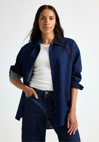Jeanskjorta i mörkblått, med krage, knappstängning och uppkavlade ärmar, lager på lager över en vit ribbad linne och mörka jeans.