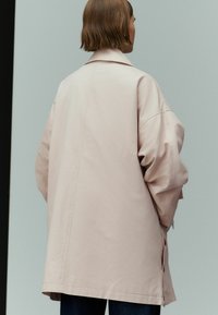 Hellrosa übergroße Jacke mit fallenden Schultern, großen Manschetten und seitlichen Knöpfen, aus glattem Stoff. Minimalistisches Design, Rückansicht.