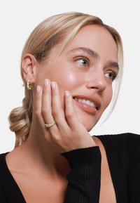 Goldene Creolen-Ohrstecker mit klaren Kristallen und einem passenden Ring mit goldenen Akzenten. Die Hand des Models ruht nahe ihrem Gesicht und zeigt den Schmuck.