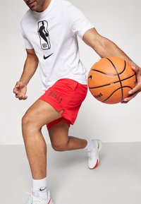 White short-sleeve T-shirt with NBA logo, paired with red mesh basketball shorts featuring "BULLS" in black. Player holds an orange basketball.