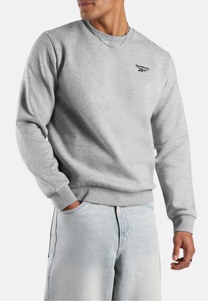 Mann trägt hellgraues Reebok-Sweatshirt und hellblaue Jeans, eine Hand in der Tasche und silberne Kettenkette.