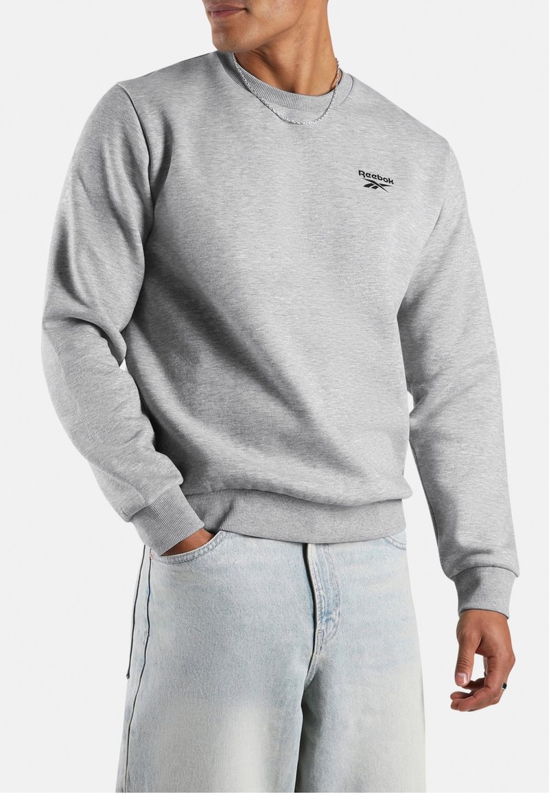 Mann trägt hellgraues Reebok-Sweatshirt und hellblaue Jeans, eine Hand in der Tasche und silberne Kettenkette.