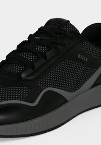 Baskets sportives BOSS noires et grises avec détails en mesh, lacets noirs et semelle texturée sur fond blanc.