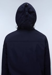 Person, die eine dunkelmarine Kapuzenjacke mit aufgesetzter Kapuze trägt, von hinten vor einem hellgrauen Hintergrund gezeigt.