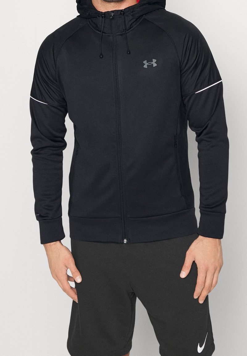 Homme portant un sweat à capuche zippé noir Under Armour et un short noir Nike, debout sur un fond uni.