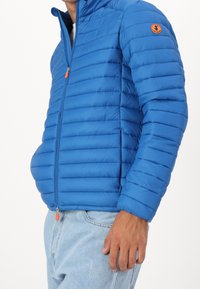 Blauwe gewatteerde jas met horizontaal quiltpatroon, frontale rits en oranje ritssluiting. Beschikt over een ronde logo patch op de linkerschouder.