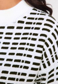 Vit stickad tröja med ett texturerat mönster av mörkgröna horisontella och vertikala ränder, med en ribbad halsringning.