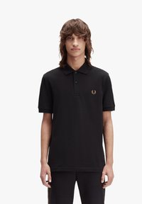 Svart bomullspolo med krage, tre knappslå, och en guld broderad logotyp på bröstet. Korta ärmar med uppvikta avslut.