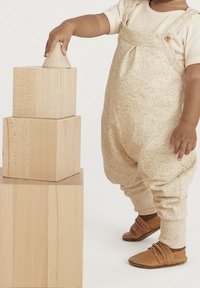 Beige, texturerade overaller på ett barn, kompletta med bruna läderskor, som staplar ljusa träblock med slät yta och skarpa kanter.