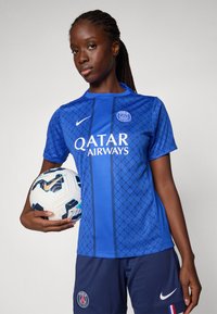 Blå atletisk tröja med geometriska mönster, vit logotyp och text. Ett par mörka shorts, håller i en mönstrad fotboll.