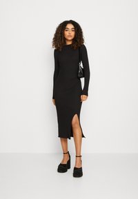 Robe midi noire côtelée à manches longues, avec une fente sur le côté. Assortie à des chaussures à plateforme noires épaisses et un petit sac à main noir.