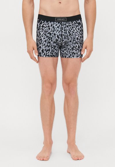 Uomo che indossa boxer a stampa leopardo nera e grigia con una cintura nera Versace, in piedi a piedi nudi su uno sfondo bianco.