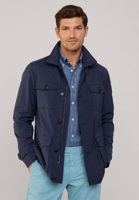 Giacca blu navy con colletto alto, tasche frontali e dettagli a bottoni, abbinata a una camicia azzurra e pantaloni chiari.