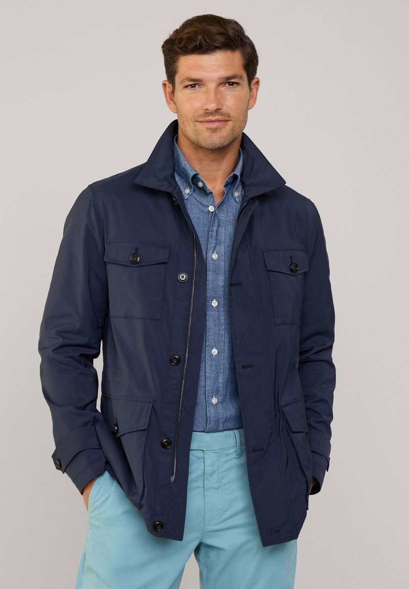 Giacca blu navy con colletto alto, tasche frontali e dettagli a bottoni, abbinata a una camicia azzurra e pantaloni chiari.