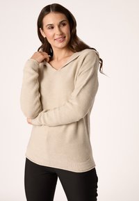 Felpa a maglia beige con scollo a V e maniche lunghe, caratterizzata da polsini e orlo inferiore a costine, abbinata a pantaloni neri attillati.
