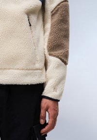 Creme fleece pullover med struktureret overflade, med brune accentlapper og en lynlås lomme. Sorte bukser synlige forneden.