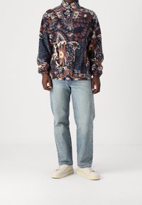 Pullover in pile con collo alto, caratterizzato da intricati motivi floreali e geometrici in blu navy, arancione e crema. Indossato con jeans di denim chiaro.