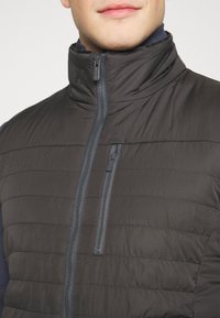 Černá péřová vesta s horizontálním prošíváním, vysokým límcem a předním zipem. Obsahuje kapsu na hrudi se zipem v kontrastní šedé barvě.