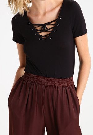 Woman wearing a black short-sleeve top with lace-up neckline and high-waisted, loose maroon pants with elastic waistband and pockets.