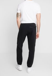 Černé fleecové tepláky s tapered střihem, elastickým pasem a manžetami na kotnících. Obsahují zipovou kapsu na zadní straně. Viditelná bílá košile a boty.