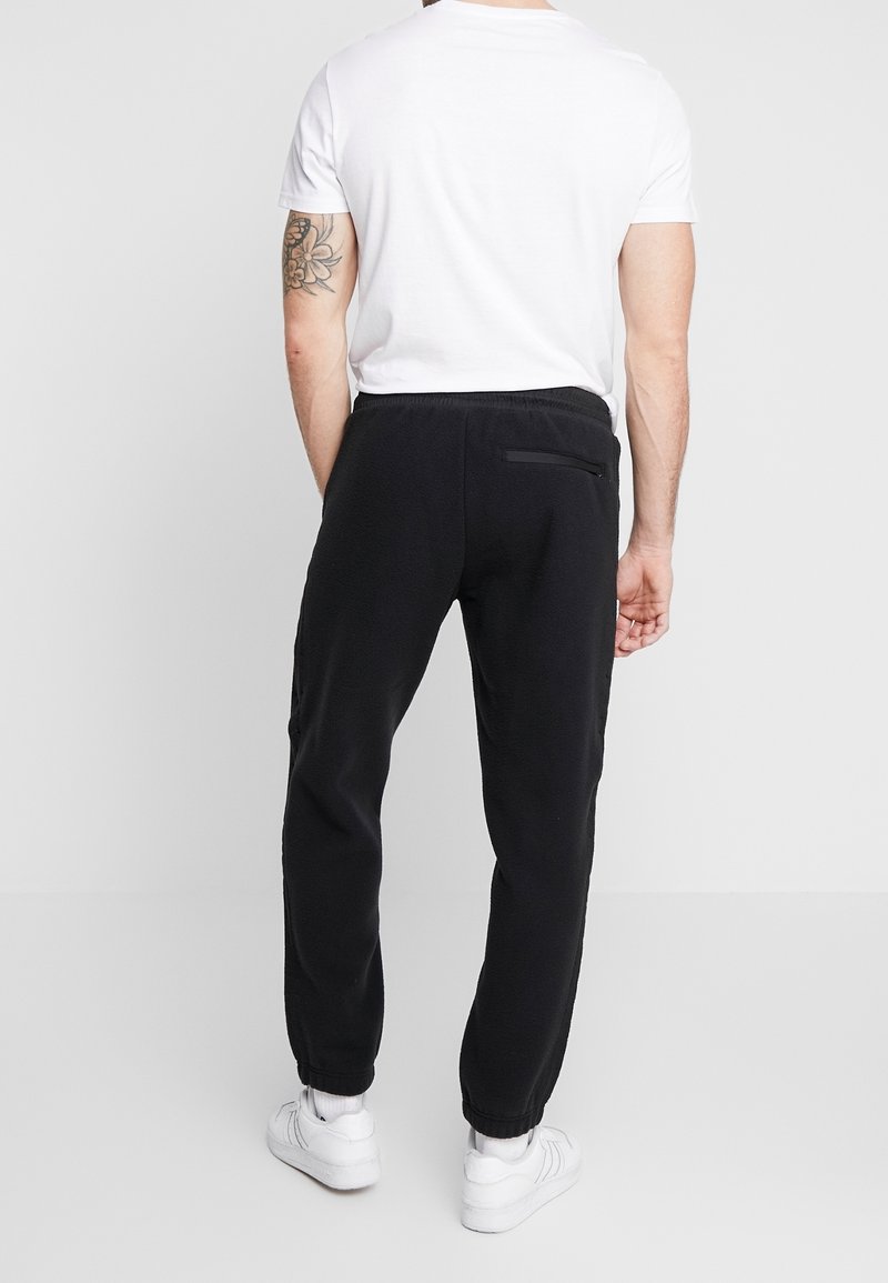 Černé fleecové tepláky s tapered střihem, elastickým pasem a manžetami na kotnících. Obsahují zipovou kapsu na zadní straně. Viditelná bílá košile a boty.