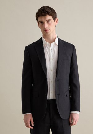 Blazer azul marino oscuro con solapas de muesca, dos bolsillos frontales y cierre de un solo botón, combinado con una camisa blanca de botones. Textura suave.