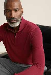 Pulover cu guler tip zip burgund în stofă texturată, asortat cu pantaloni gri. Fundalul prezintă o canapea închisă la culoare.