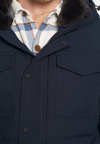 Hombre vestido con una chaqueta azul marino con bolsillos en el pecho abotonados y una camisa de cuadros en azul, blanco y marrón debajo, parcialmente cerrada.