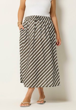 Frau trägt einen diagonal gestreiften braun-weißen Midi-Rock mit Kordelzug in der Taille, weißes Tanktop, Goldarmband und weiße Sandalen.
