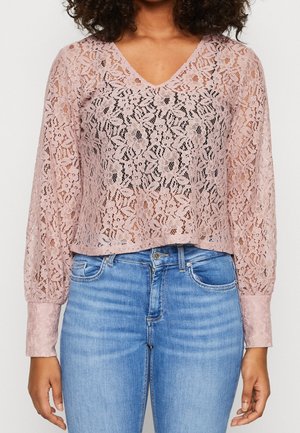 Frau trägt ein langärmeliges, rosa, florales Spitzenoberteil mit V-Ausschnitt und blaue, hoch taillierte Jeans, Hände entspannt an den Seiten.