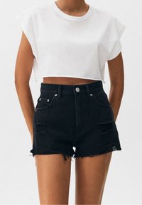 Camiseta blanca corta con mangas enrolladas combinada con pantalones cortos de mezclilla negros de tiro alto con detalles desgastados y dobladillo deshilachado.