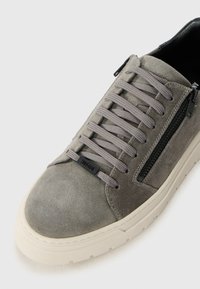 Grå semsket sneakers med grå snøringer, sort sideglidelås, hvit gummisål, og "MORATO" merke-tag på en hvit bakgrunn.