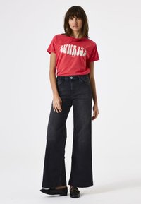 Rotes T-Shirt mit dem Text "SUNRISE" und kurzen Ärmeln, kombiniert mit hochgeschnittenen, schwarzen, weit geschnittenen Jeans; einfache schwarze Ballerinas runden das Outfit ab.