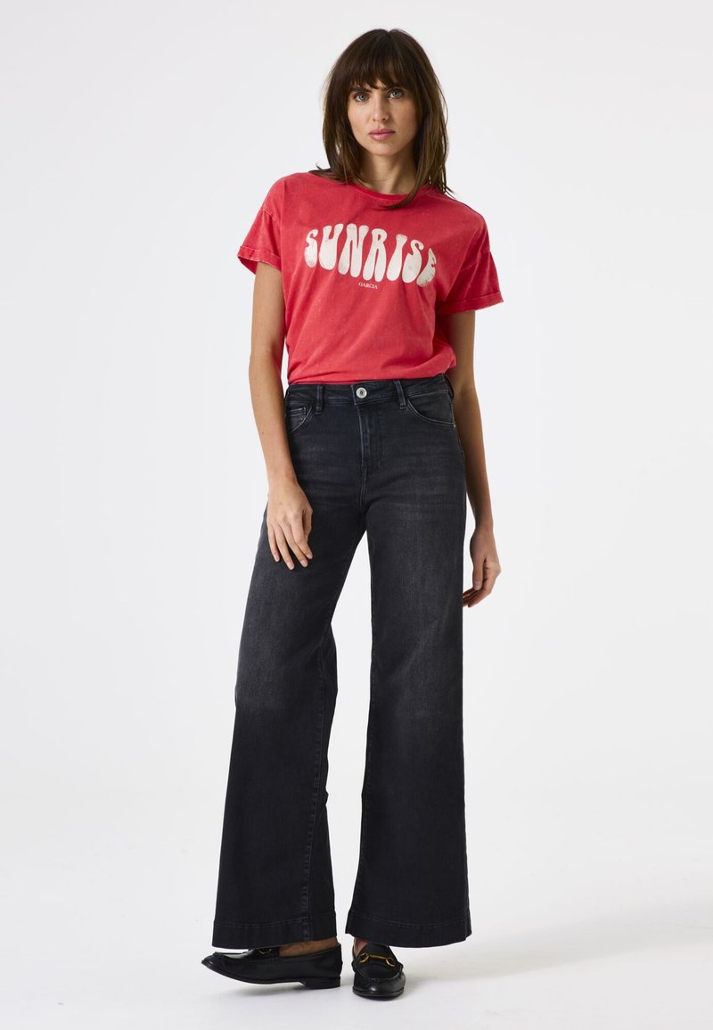 Rotes T-Shirt mit dem Text "SUNRISE" und kurzen Ärmeln, kombiniert mit hochgeschnittenen, schwarzen, weit geschnittenen Jeans; einfache schwarze Ballerinas runden das Outfit ab.