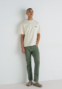 Cream cotton t-shirt with a small blue graphic on the left chest, paired with olive green pants and tan slip-on shoes. Standing against a white wall.