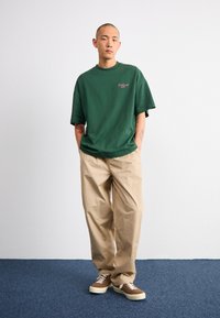 Camiseta de algodón verde con mangas cortas y escote redondo, con un pequeño logo rojo. Combinada con pantalones beige de corte holgado y zapatillas marrones.