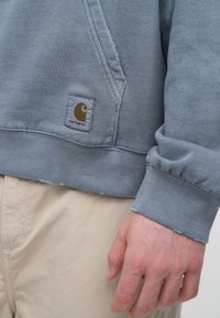 Szary pulower z kapturem wykonany z miękkiej tkaniny, wyposażony w boczną kieszeń i ściągacze na mankietach. Na przodzie znajduje się charakterystyczna naszywka z logo Carhartt w żółtym i białym kolorze.